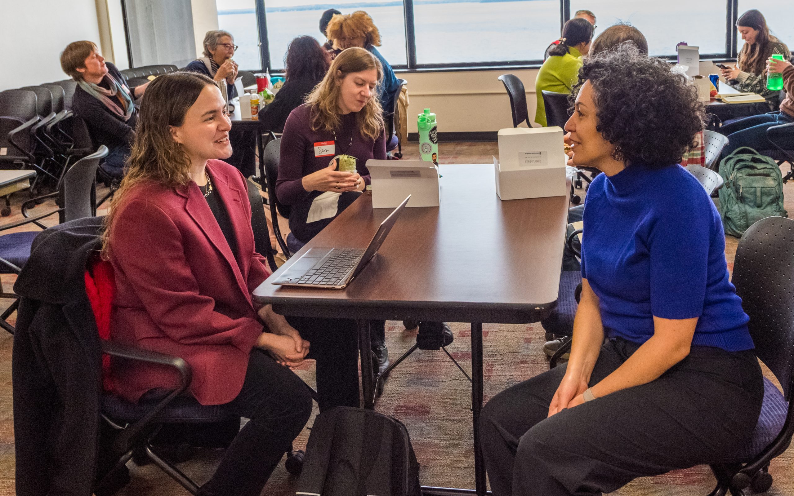This image features two women sitting across from each other at a table. The one to the right is a Black woman with a cropped and curly haircut wearing a dark blue, short-sleeved sweater and black jeans. The woman to the left is a white women with medium length black and brown hair, wearing a maroon suit jacket with black pants. 