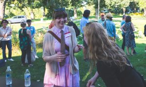 A white student wearing a pink dress with a brown tie and a brown sweater smiles while in conversation with someone facing away from the camera. The featured student hair is pulled back into a ponytail, and she has eyebrow length bangs.